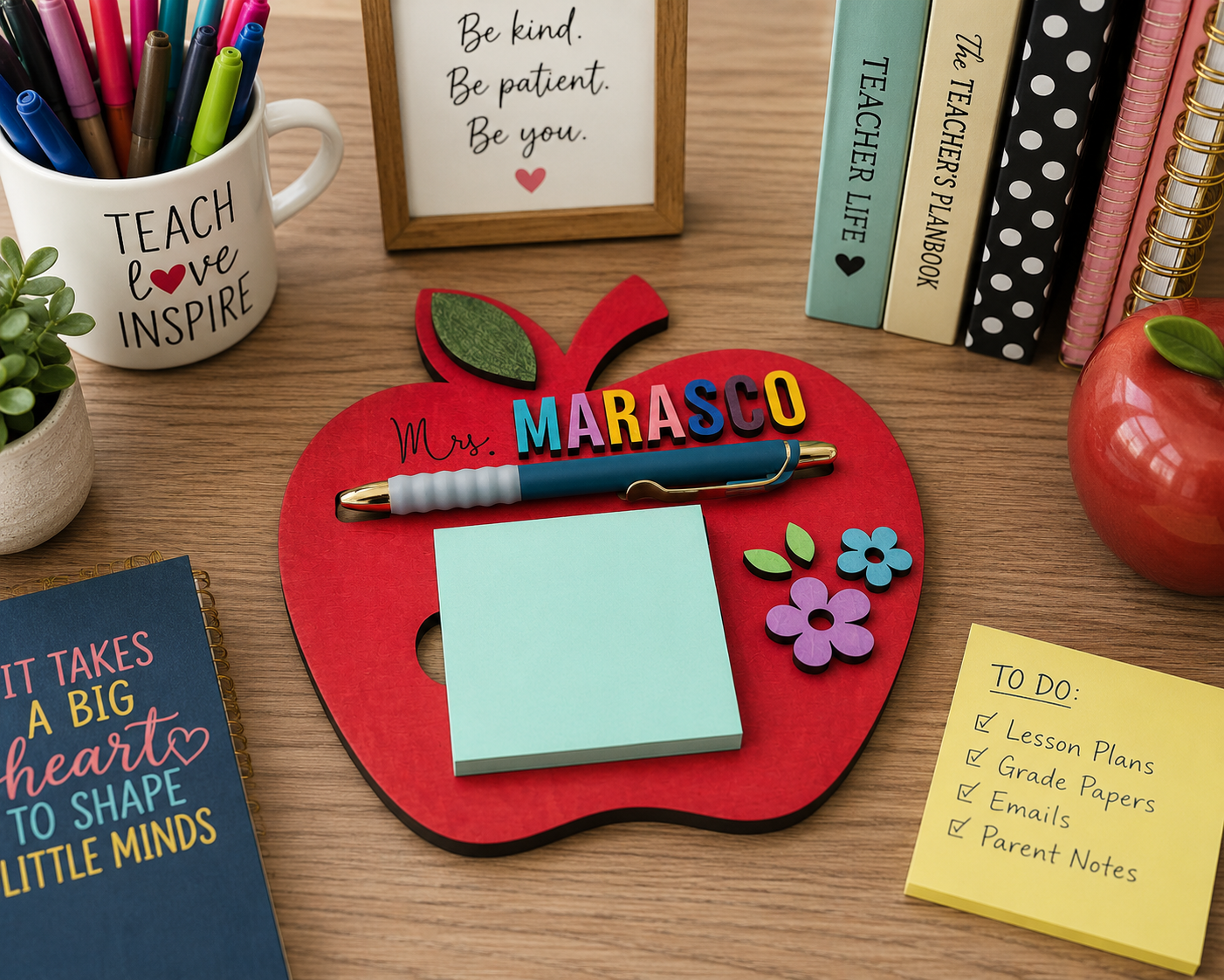 A red apple-shaped desk organizer with a personalized name 'MARASCO' in colorful letters, holding a blue pen and a light blue sticky note pad, styled on a wooden desk with school supplies and books.